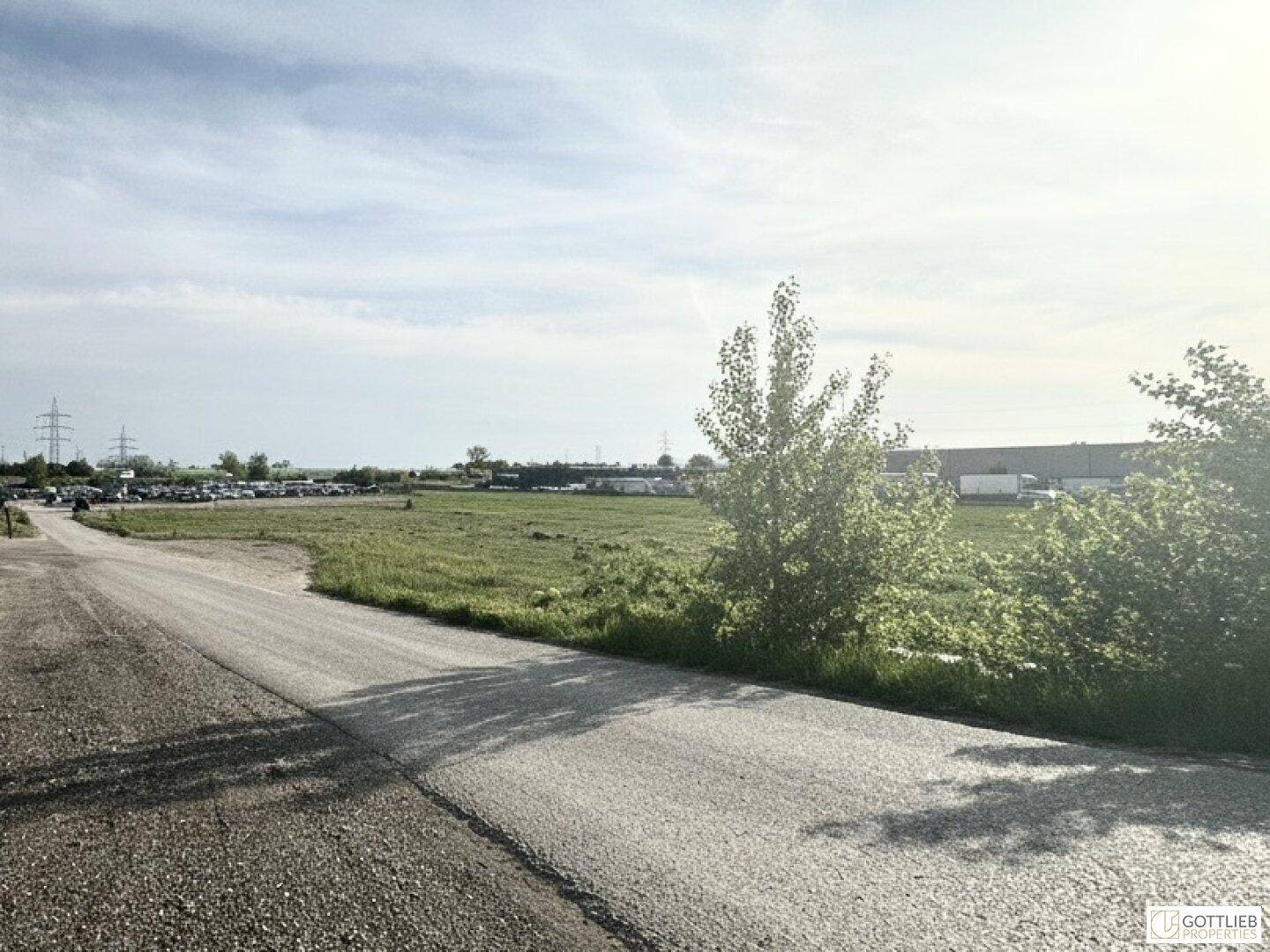 Offenes Gelände mit Zufahrtsstraße, grünen Flächen und einem Parkplatz im Hintergrund unter hellem Himmel.