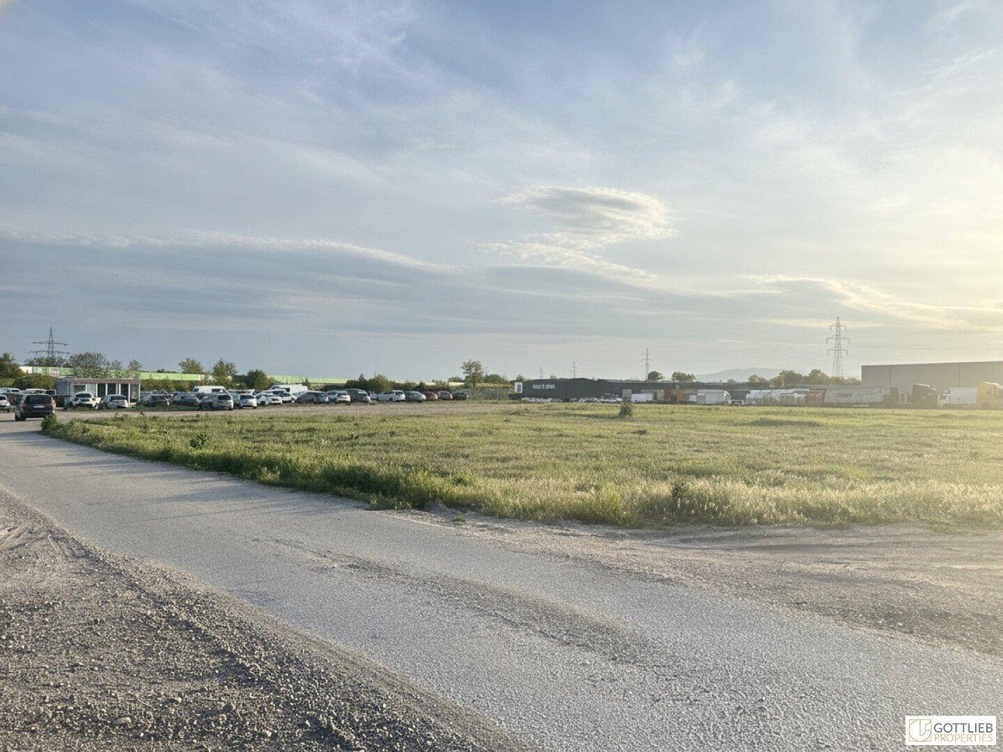Weitläufiges Gelände mit Zufahrtsweg, grünen Wiesen und einem großen Parkplatz im Hintergrund bei Sonnenuntergang.
