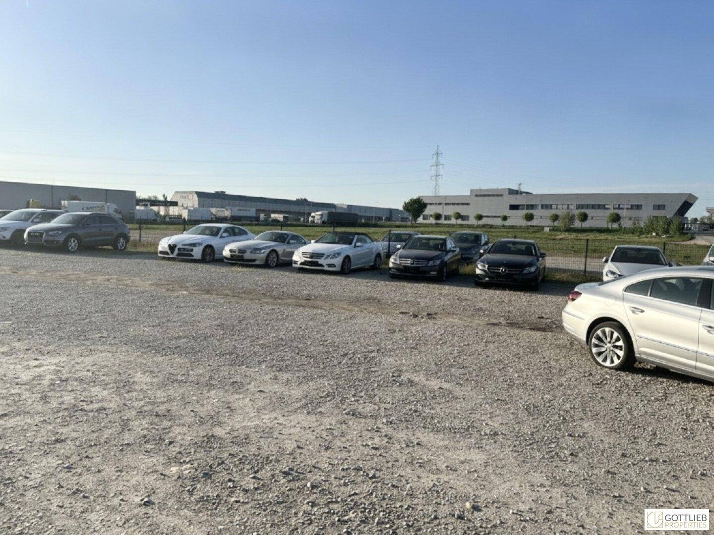 Schotterparkplatz mit einer Reihe geparkter Fahrzeuge und Industriegebäuden in der Ferne unter klarem Himmel.