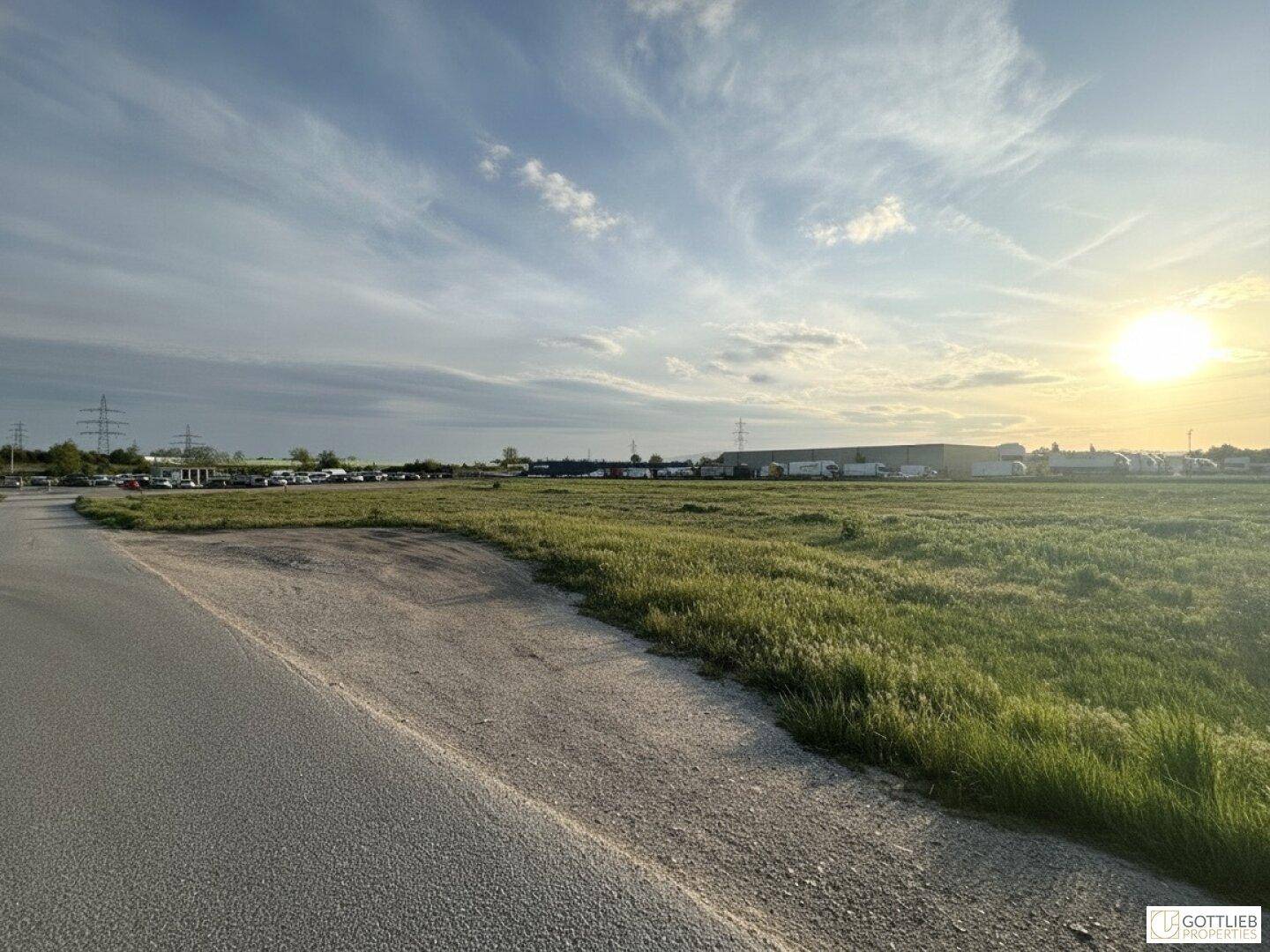 Weitläufiges Gelände mit Zufahrtsweg, grünen Wiesen und einem großen Parkplatz im Hintergrund bei Sonnenuntergang.