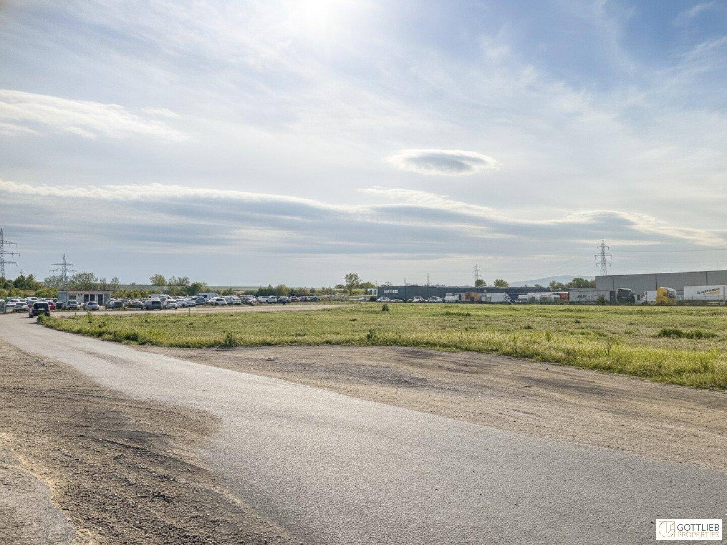 Weitläufiges Außengelände mit Zufahrtsstraße, Grünflächen und Parkplätzen im Hintergrund unter bewölktem Himmel.