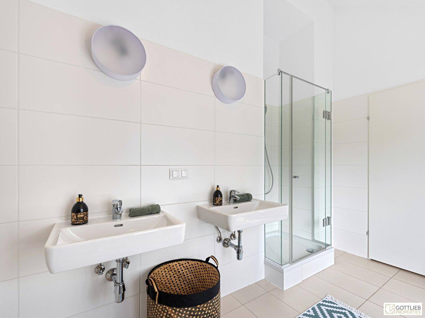 Modernes Badezimmer mit zwei Waschbecken, einer Glasdusche und hellen, großformatigen Fliesen.