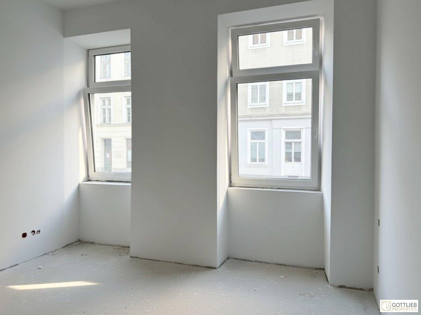 Leerer Raum mit weißen Wänden, Betonboden und zwei hohen Fenstern mit Blick auf die Umgebung.