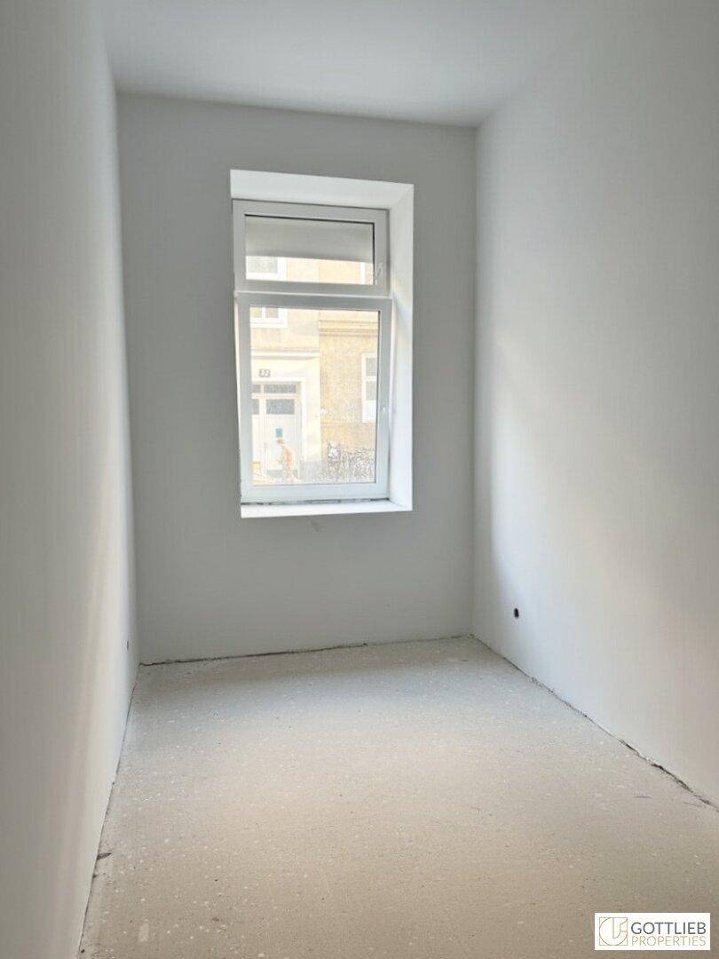 Kleiner, heller Raum mit einem Fenster, weißen Wänden und einem einfachen Betonboden.