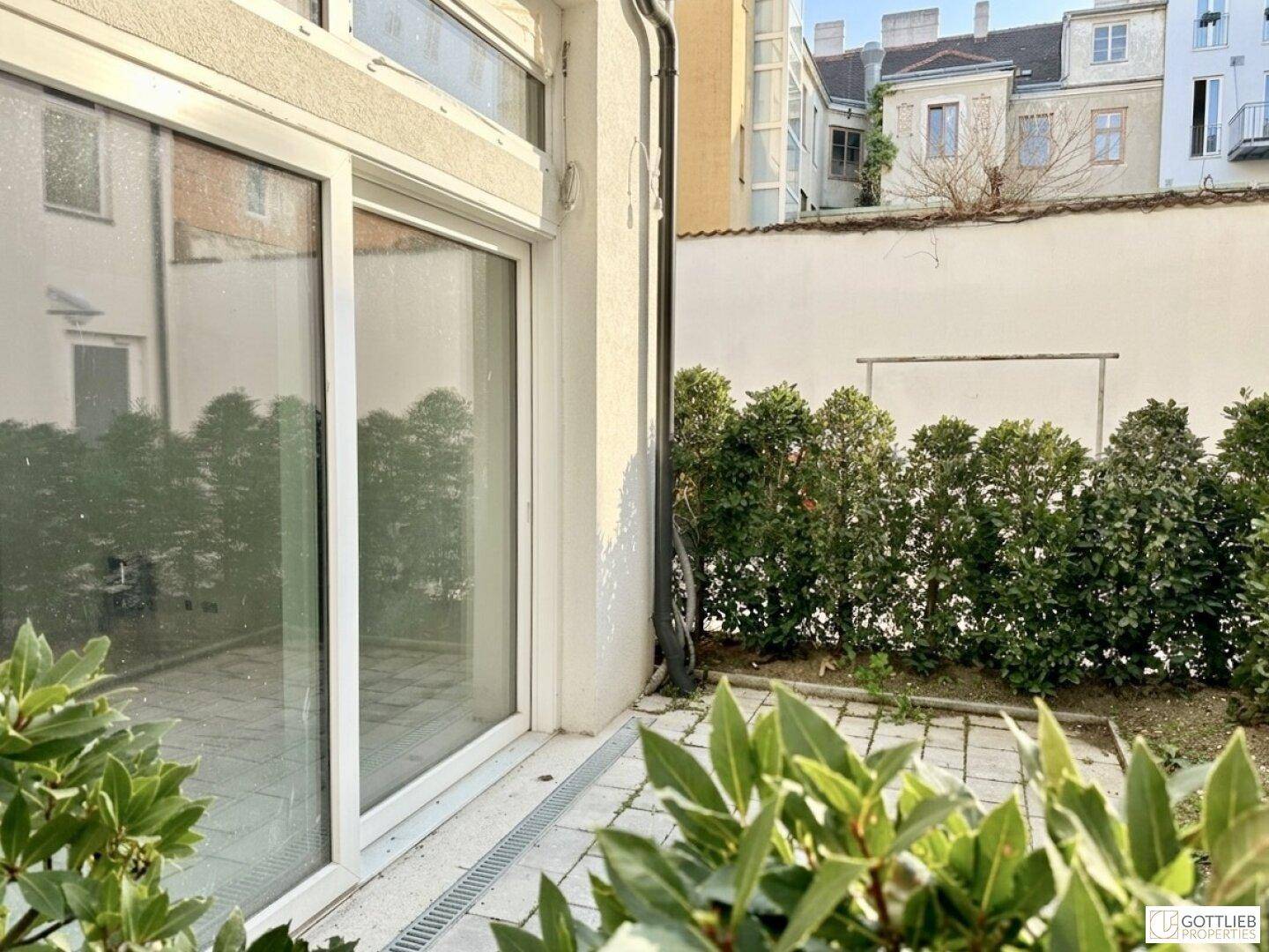 Große Glasschiebetür öffnet sich zu einer kleinen gepflasterten Terrasse mit grünen Hecken.