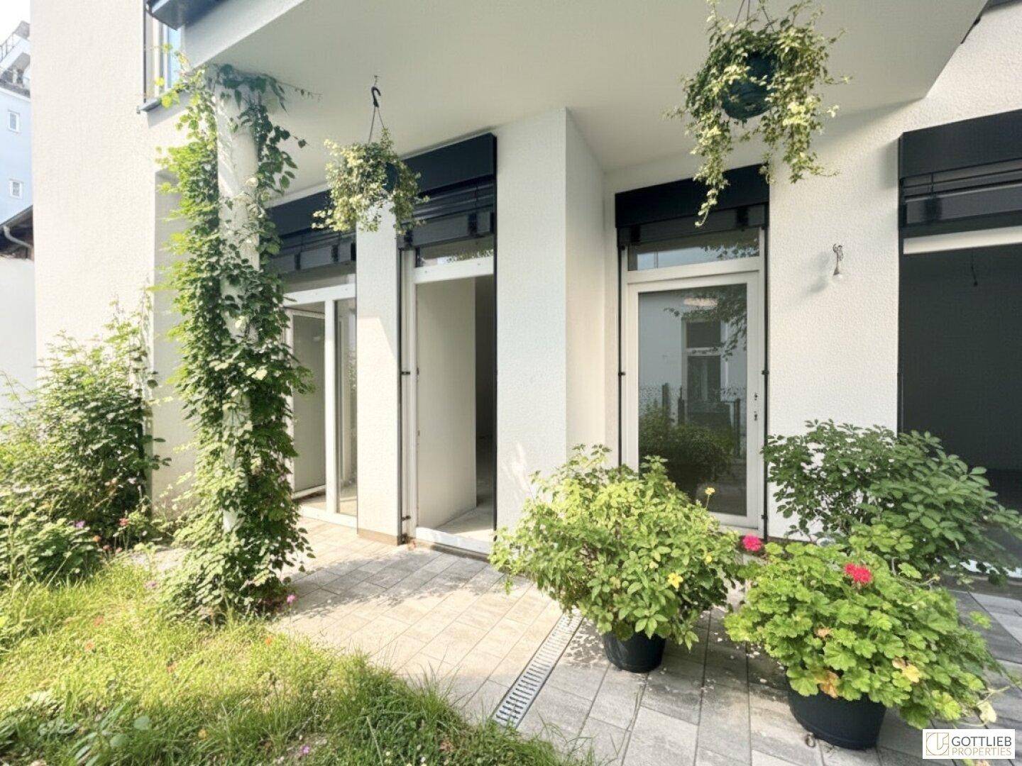 Sonniger Terrassenbereich mit üppiger Bepflanzung und hängenden Blumenampeln am Gebäude.