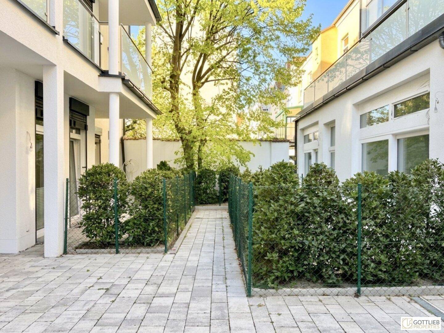 Gepflasterter Weg zwischen Gebäuden, gesäumt von Hecken und Bäumen in einem Innenhof.