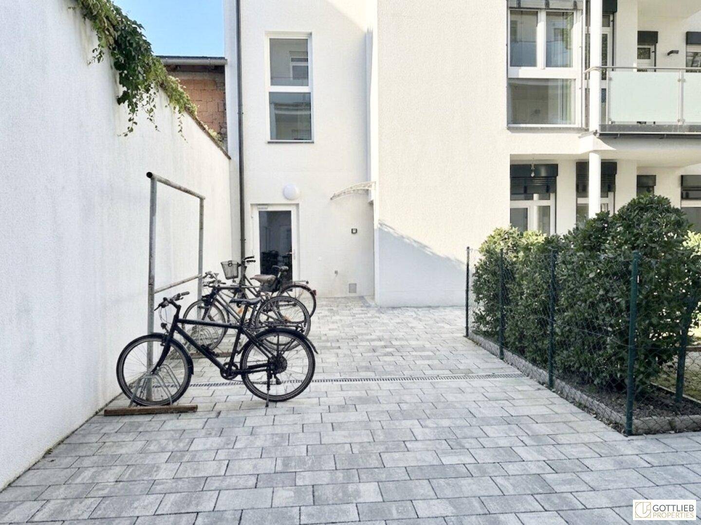 Gepflasterter Bereich mit Fahrradständern und Fahrrädern vor einer modernen, weißen Gebäudefassade.