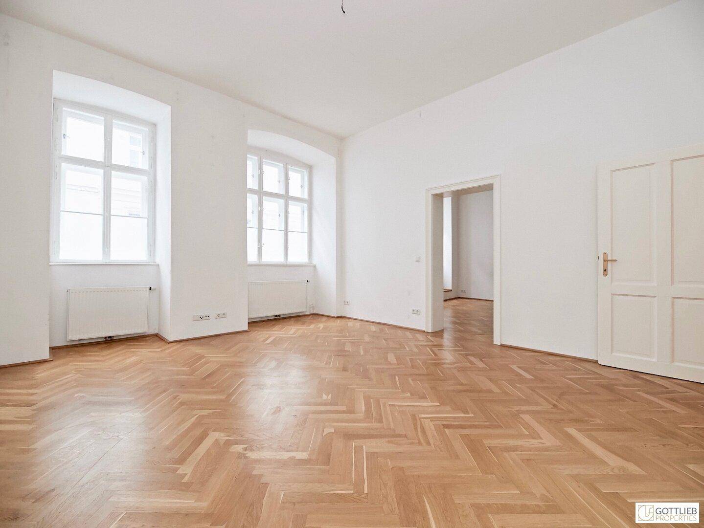 Geräumiges Wohnzimmer mit Fischgrätparkett, hohen Decken und großen Fenstern, die viel Licht hereinlassen.