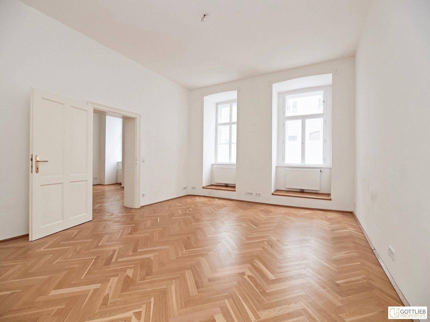 Geräumiges Zimmer mit Fischgrätparkett, hohen Fenstern und einer Doppeltür, die zu einem weiteren Raum führt.