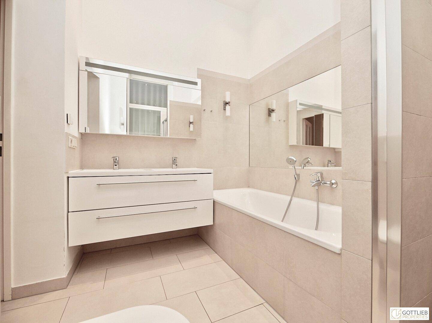Modernes Badezimmer mit Doppelwaschbecken, Badewanne und großen Spiegeln, hell gefliest.