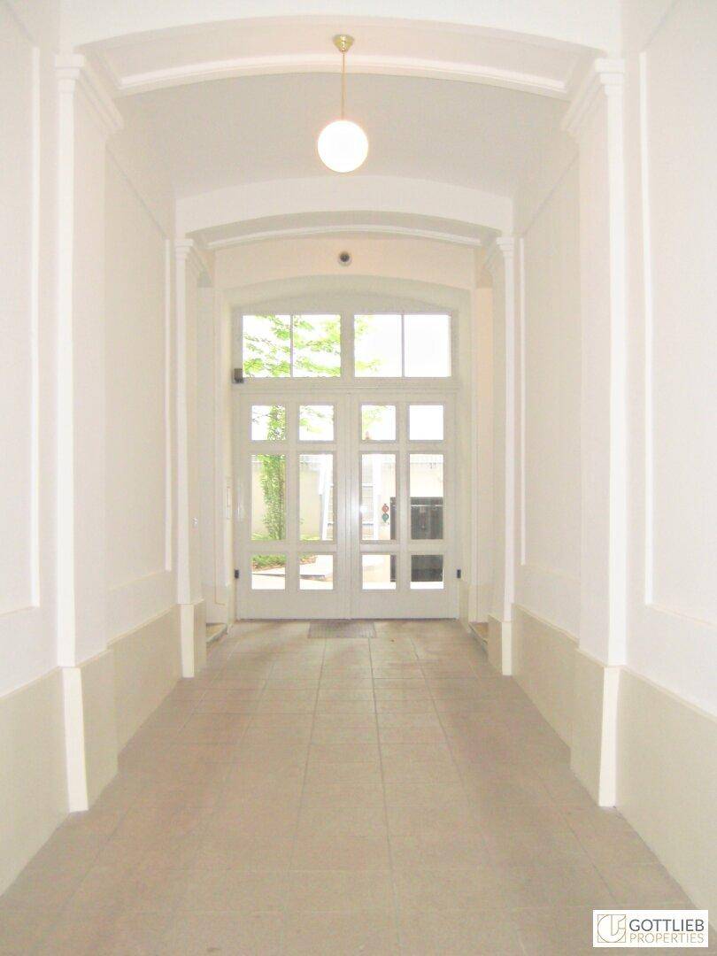 Heller Flur mit hohen Decken, Fliesenboden und einer großen Glastür am Ende.