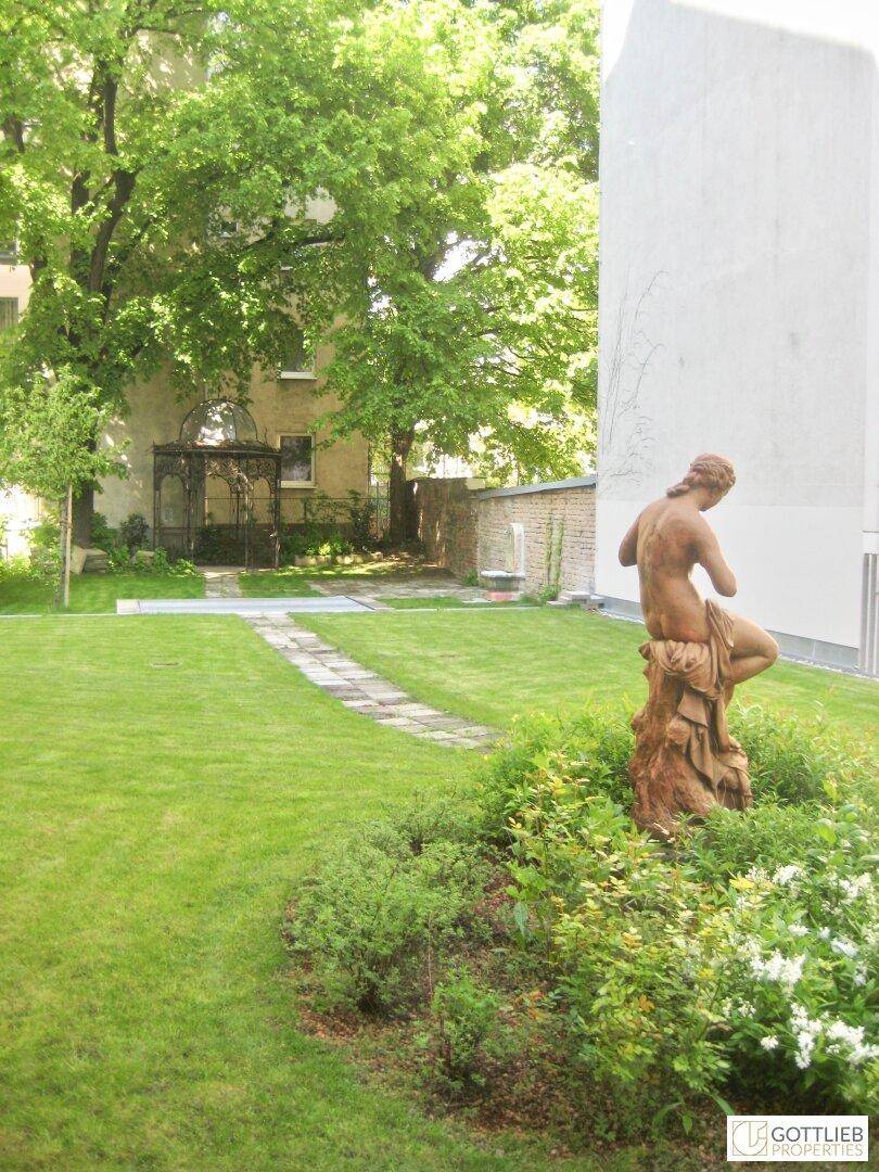 Gartenbereich mit Rasen, Büschen und einer Statue, im Hintergrund ist ein Gebäude zu sehen.