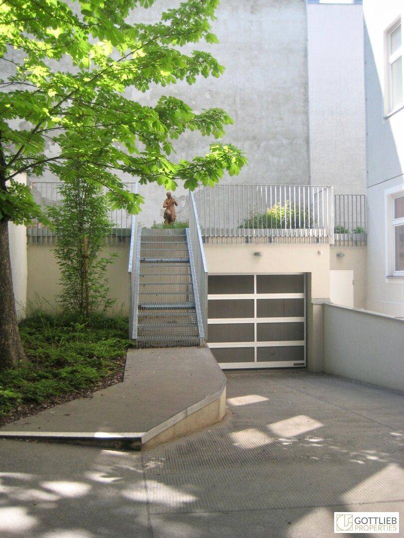 Außenbereich mit Garagenzufahrt, Treppe und einer Statue im Grünen, umgeben von Gebäuden.