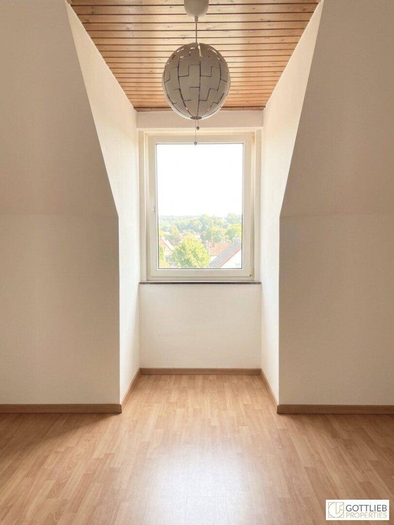 Dachgeschossraum mit Holzdecke, Laminatboden und einer modernen Deckenleuchte über dem Fenster.
