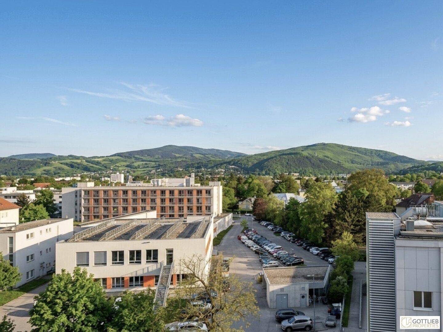 Weitläufiger Blick über die Stadtlandschaft mit grünen Bergen am Horizont unter blauem Himmel.