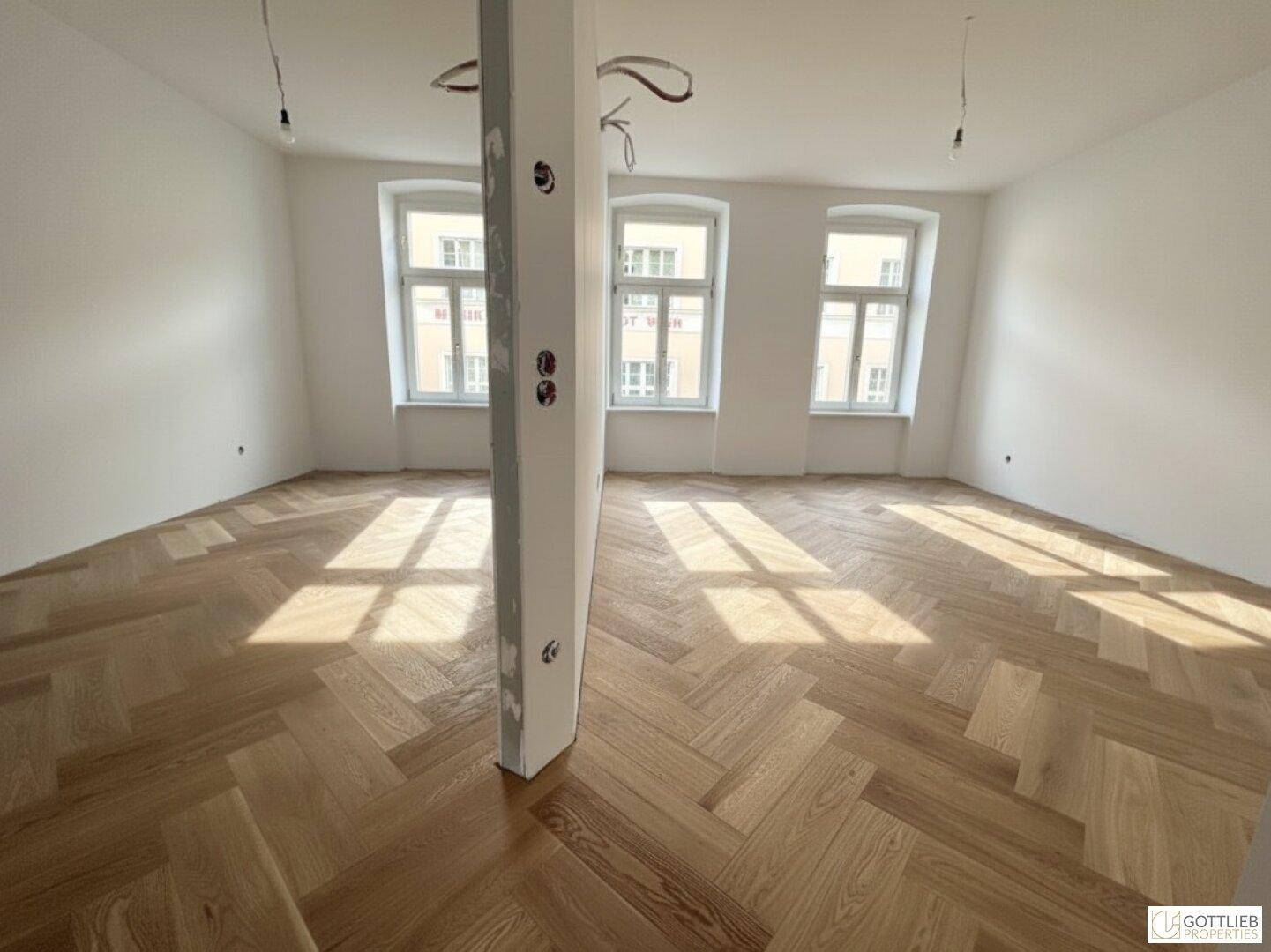 Großzügiger, heller Raum mit Fischgrätparkett und mehreren Fenstern, ideal für offenes Wohnen.