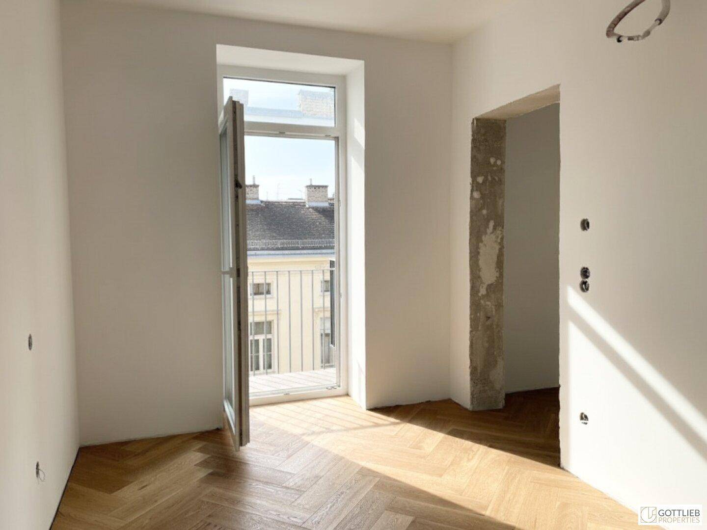 Sonniger Raum mit Fischgrätparkett und offenem Fenster, bietet Zugang zu einem französischen Balkon.