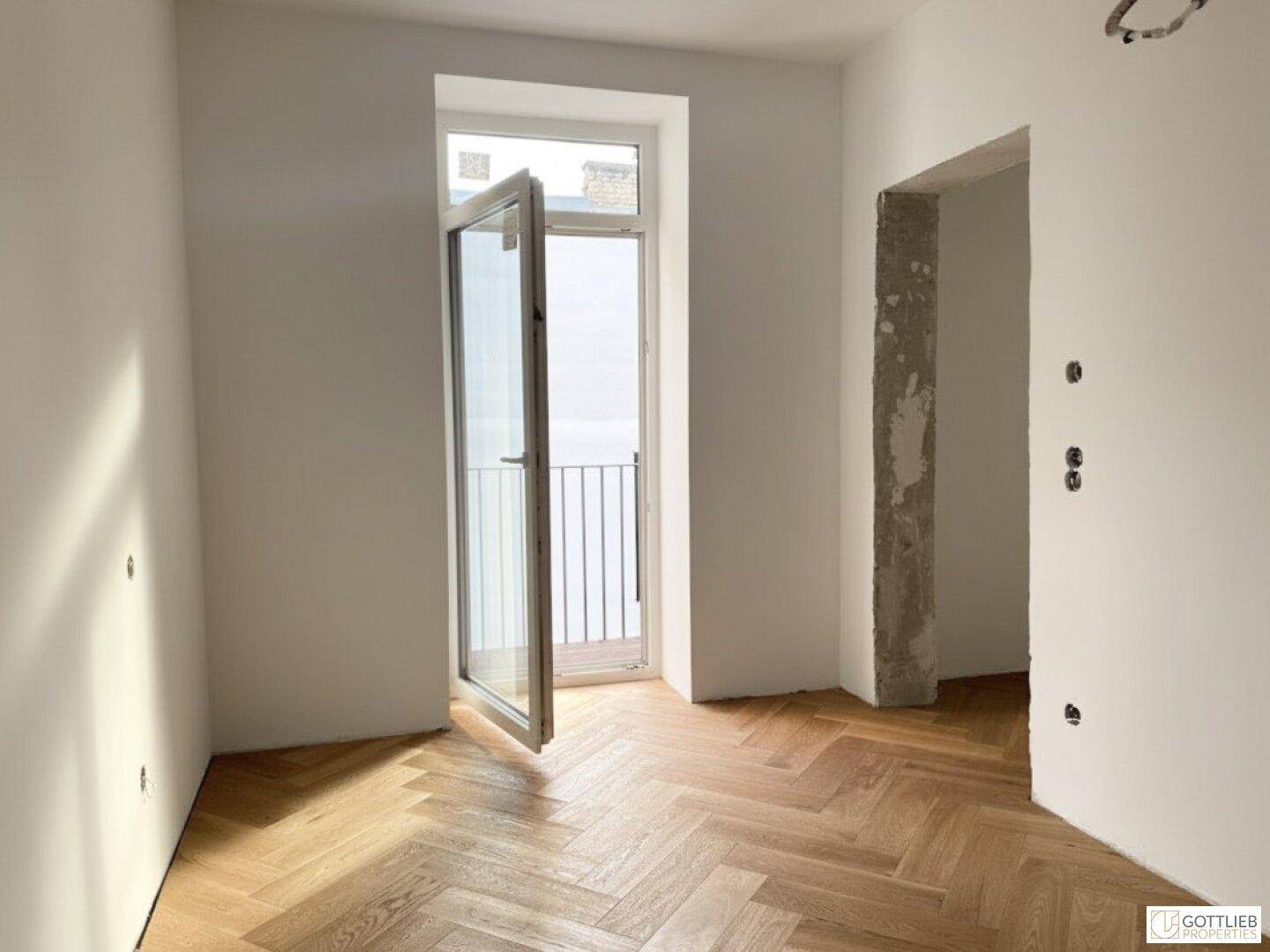 Heller Raum mit Fischgrätparkett und Zugang zu einem französischen Balkon, mit Blick nach draußen.