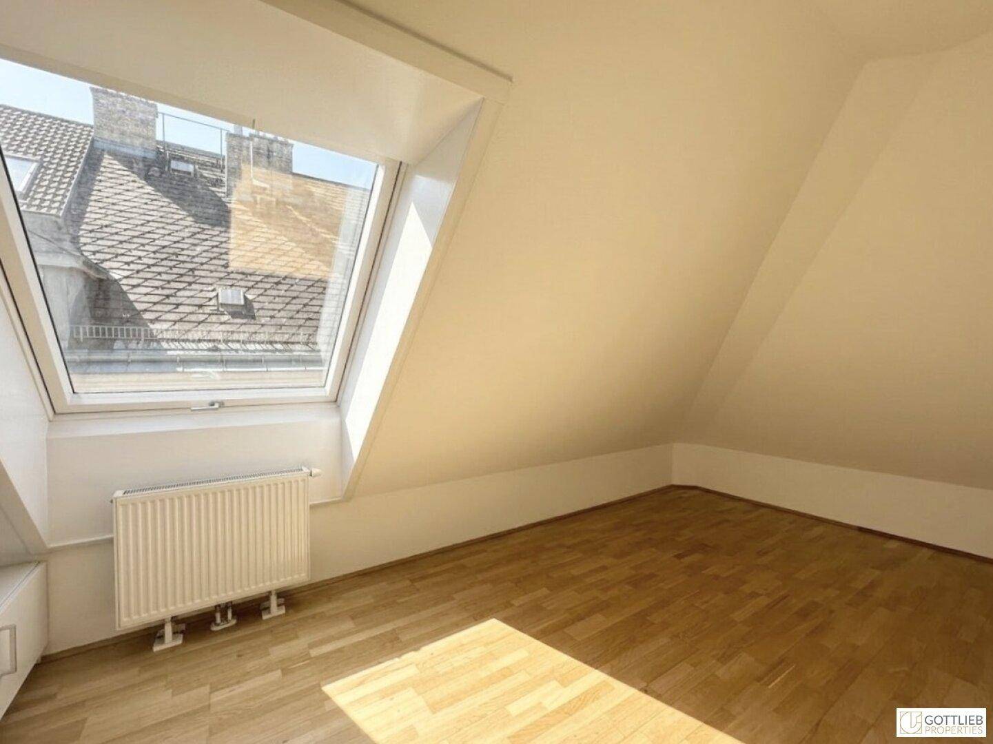 Helles Dachgeschosszimmer mit Holzboden und Dachfenster, das einen Blick auf die Dächer bietet.