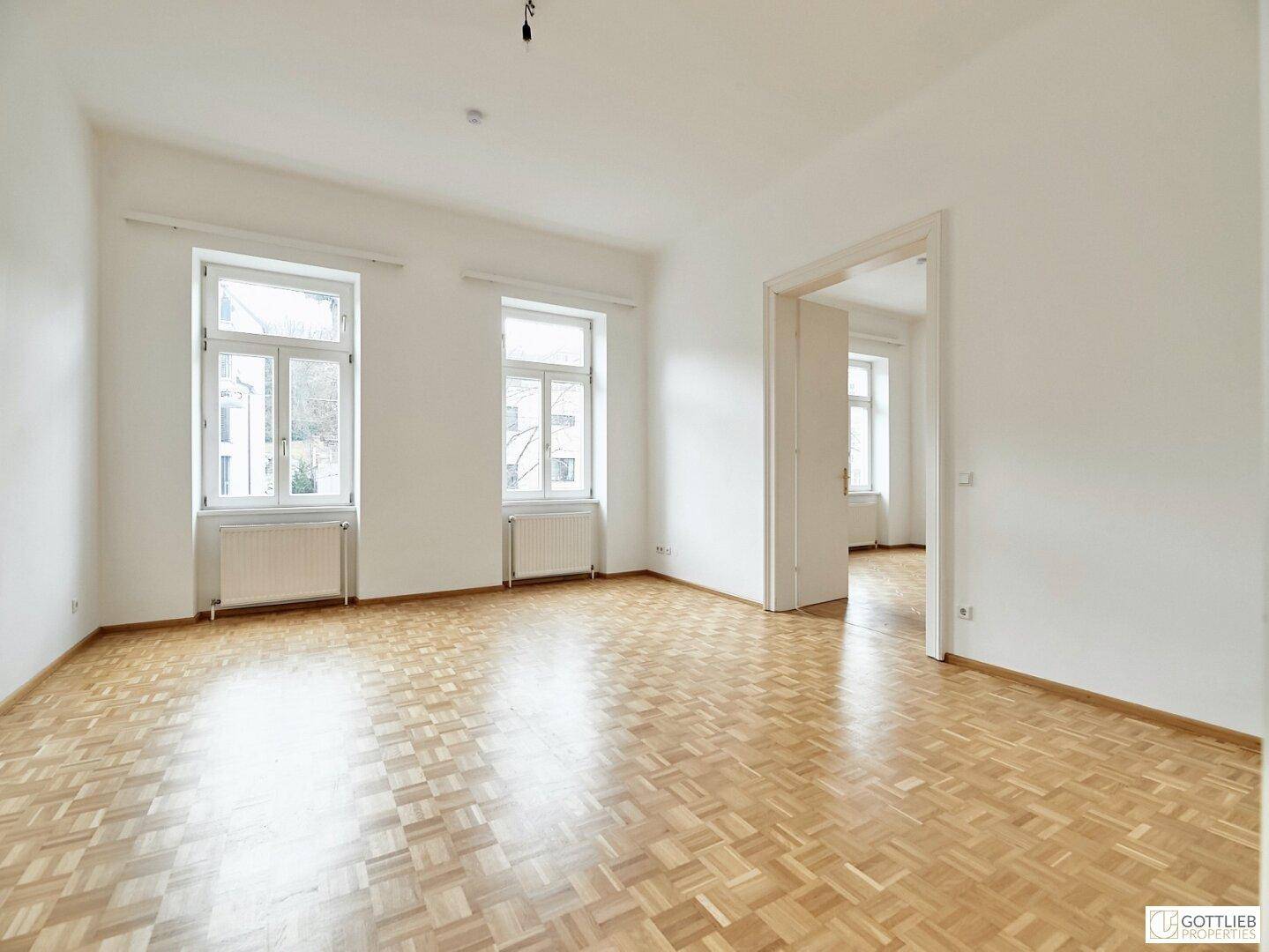 Geräumiges, helles Zimmer mit klassischem Parkettboden und zwei großen Fenstern, ideal für Wohnzwecke.