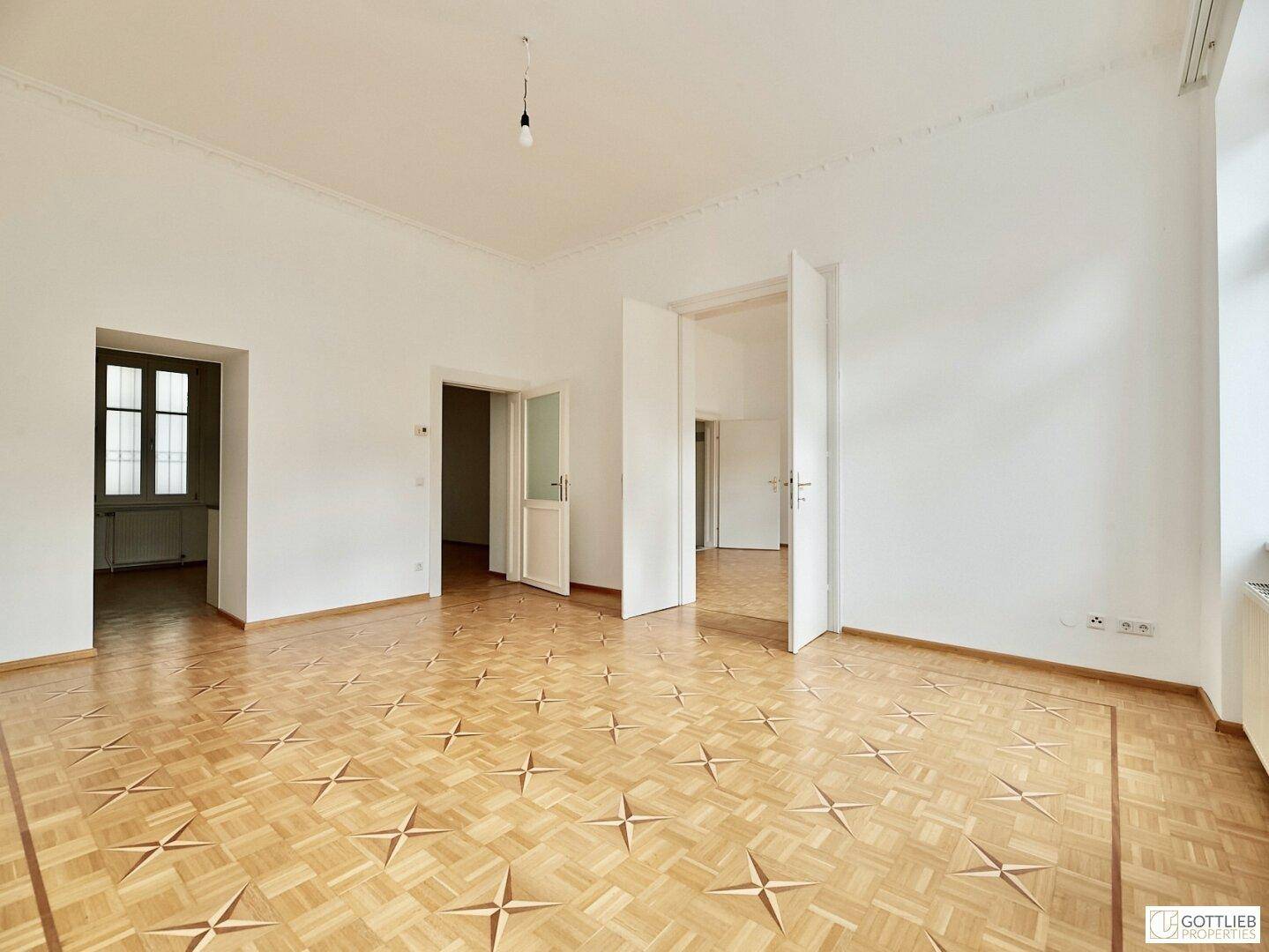 Großes, helles Zimmer mit Parkettboden und hohen Decken, verbunden durch mehrere Türen zu anderen Räumen.
