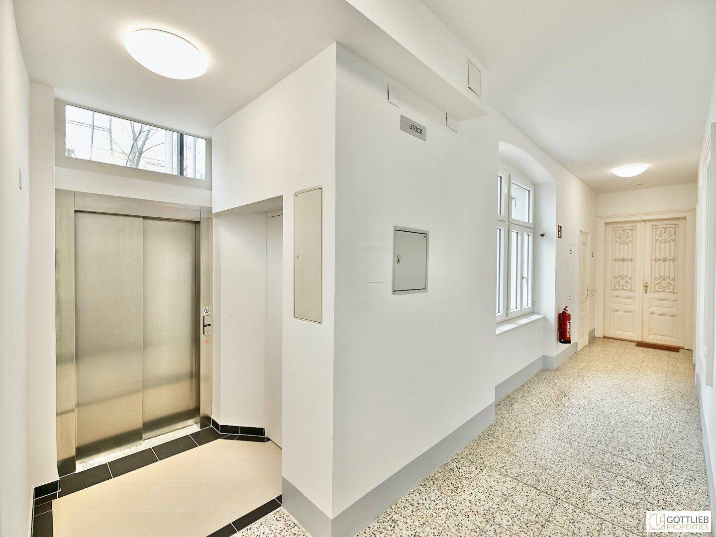Gemeinschaftsflur mit Terrazzoboden, Aufzug und natürlichem Lichteinfall durch ein Fenster.