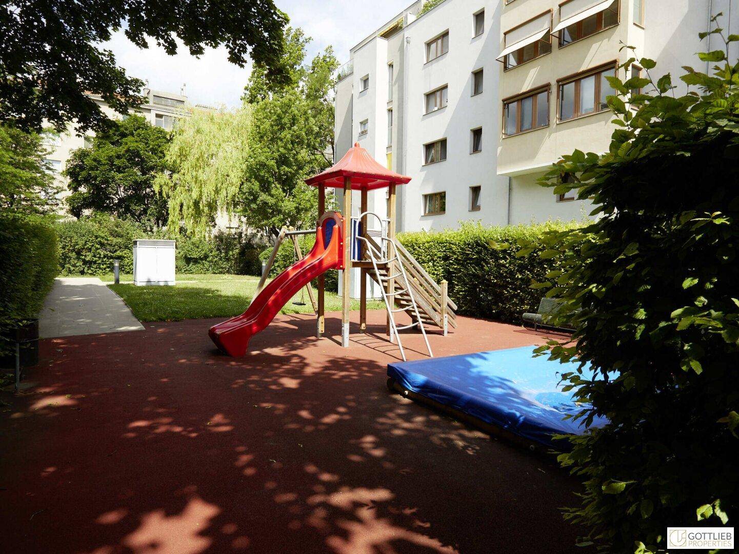 Kinderspielplatz mit roter Rutsche und Klettergerüst, umgeben von grünen Hecken in der Wohnanlage.