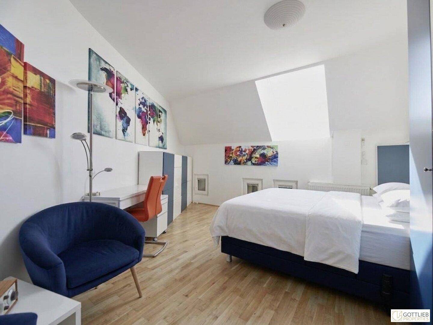 Helles Schlafzimmer mit Bett, Schreibtisch und farbenfroher Wandkunst unter dem Dachfenster.
