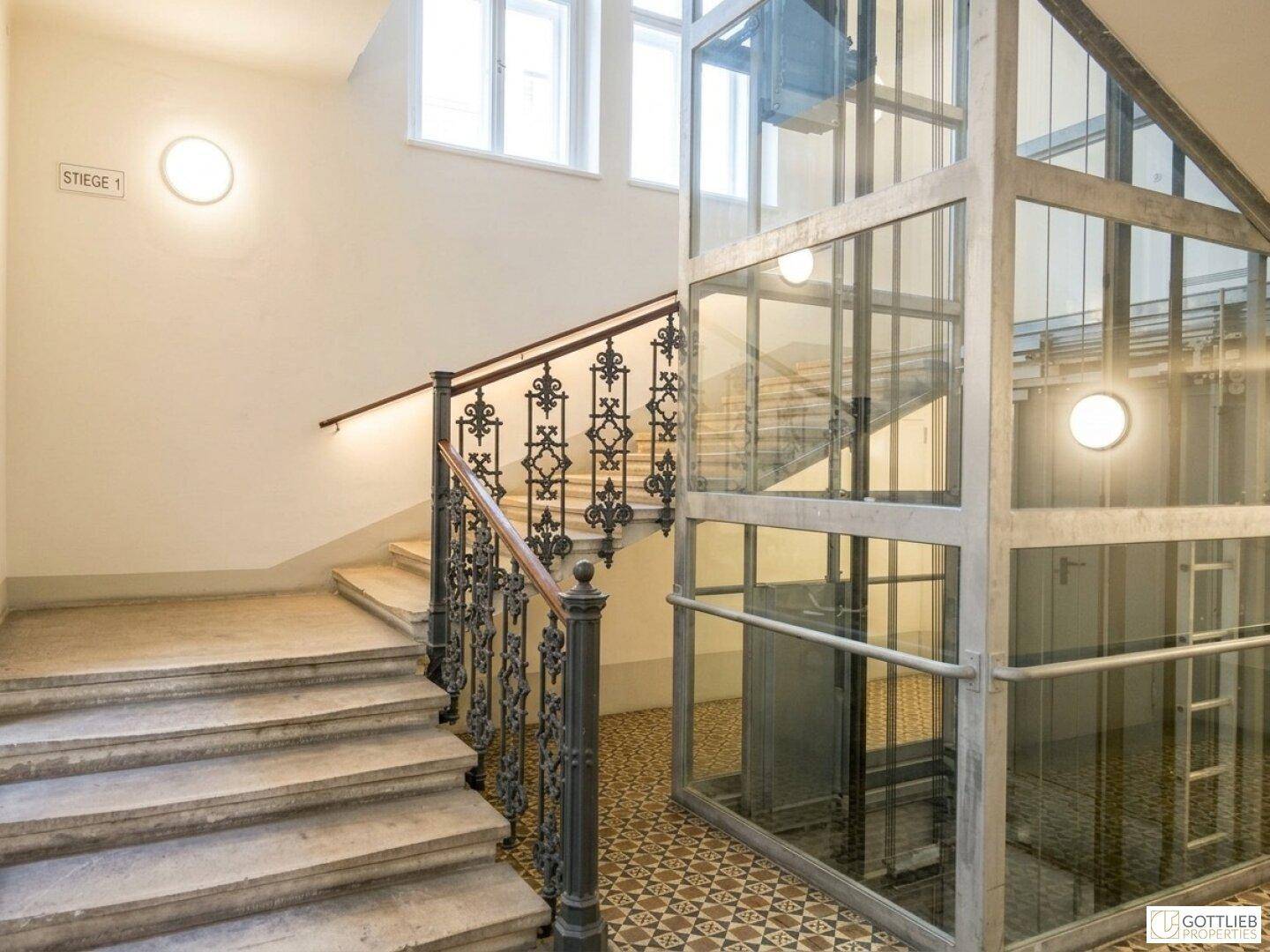 Historisches Treppenhaus mit schmiedeeisernem Geländer und modernem Glasaufzug.