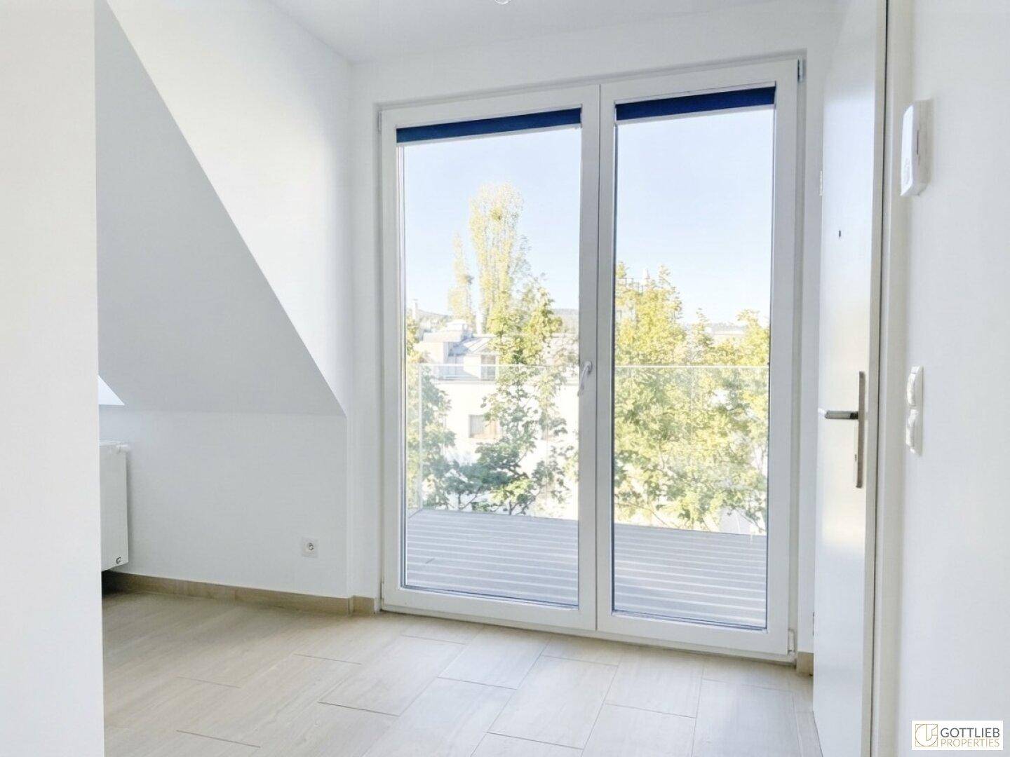 Helles Zimmer mit Fliesenboden, Dachschräge und großen Fenstertüren zum Balkon.