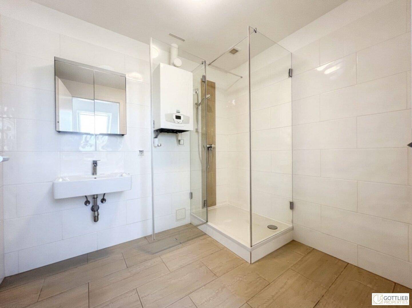 Helles Badezimmer mit Duschkabine, Waschbecken und Warmwasserbereiter an der Wand.