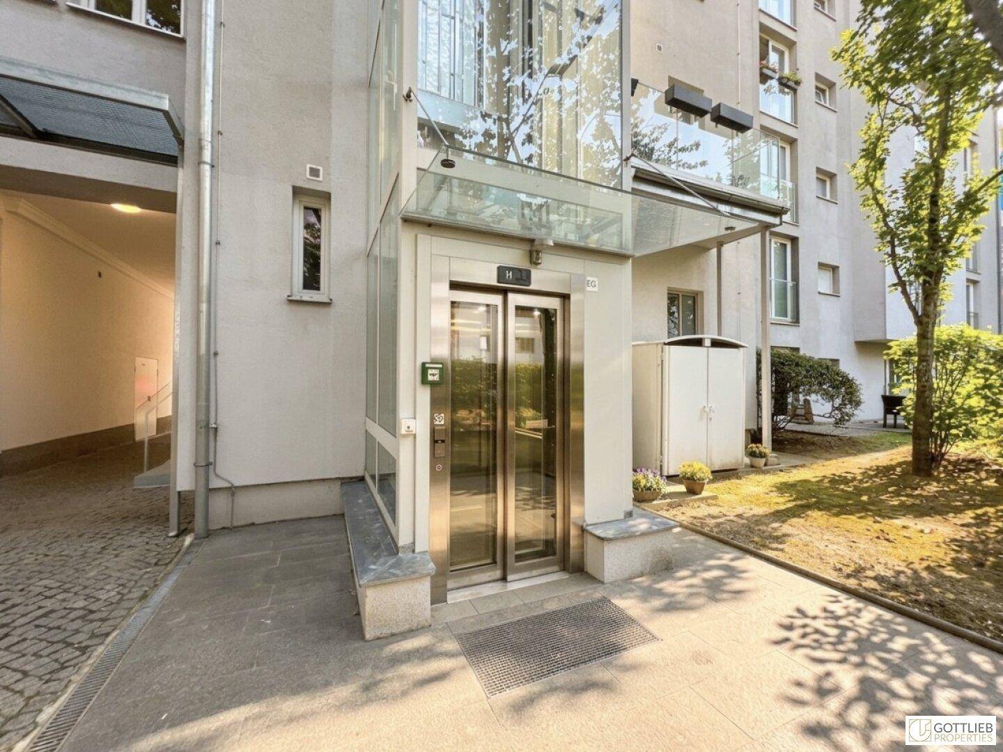 Modernes Gebäude mit gläsernem Aufzug am Eingangsbereich und gepflegter Außenanlage.
