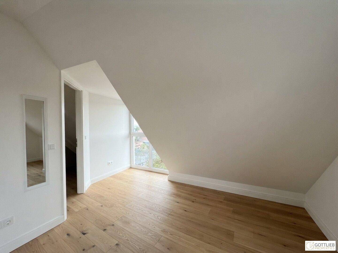 Leeres Zimmer im Dachgeschoss mit Holzboden, schrägen Wänden und einem großen Fenster.