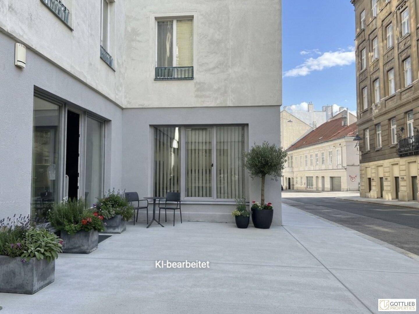 Gebäudefassade mit kleiner Terrasse und Sitzgelegenheiten, Blick auf die Straße und umliegende Gebäude.