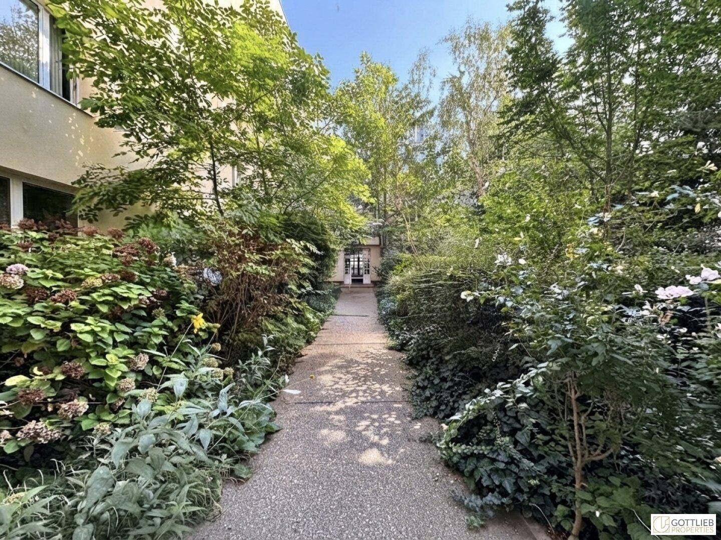 Grüner, bewachsener Gartenweg führt zu einem Gebäudeeingang, umgeben von üppiger Vegetation.
