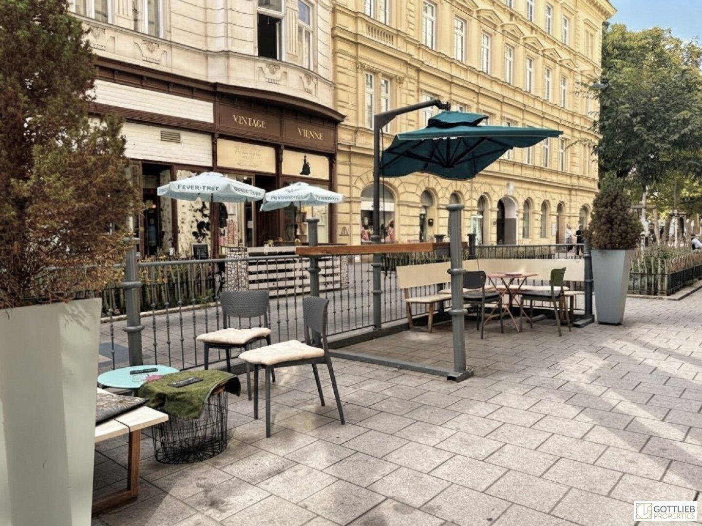 Einladender Außenbereich mit Tischen, Stühlen und Sonnenschirmen vor einer historischen Ladenfassade.