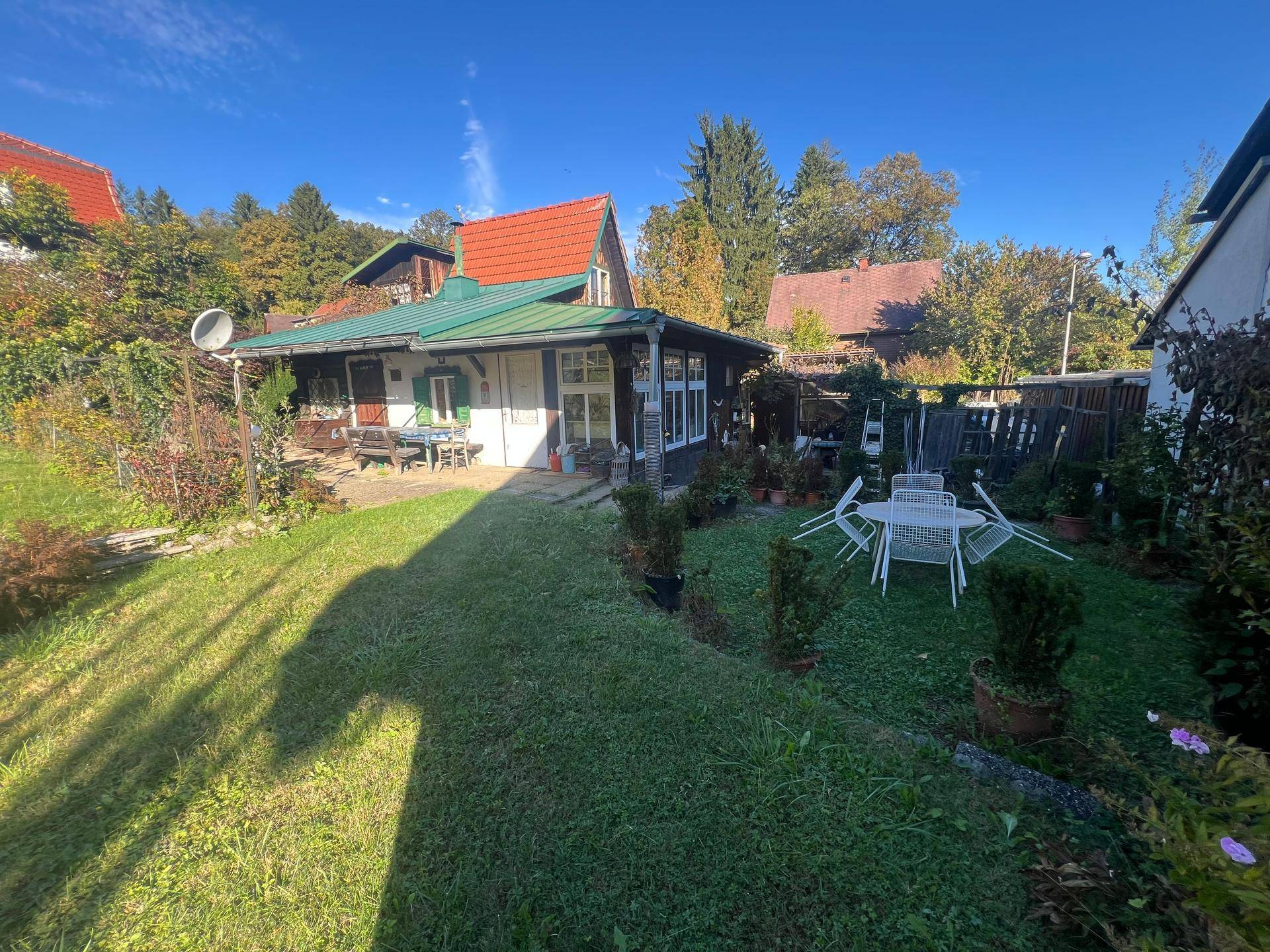 Idyllischer Garten mit zwei möblierten Sitzbereichen und einem charmanten Haus mit grünem Dach, umgeben von viel Grün.