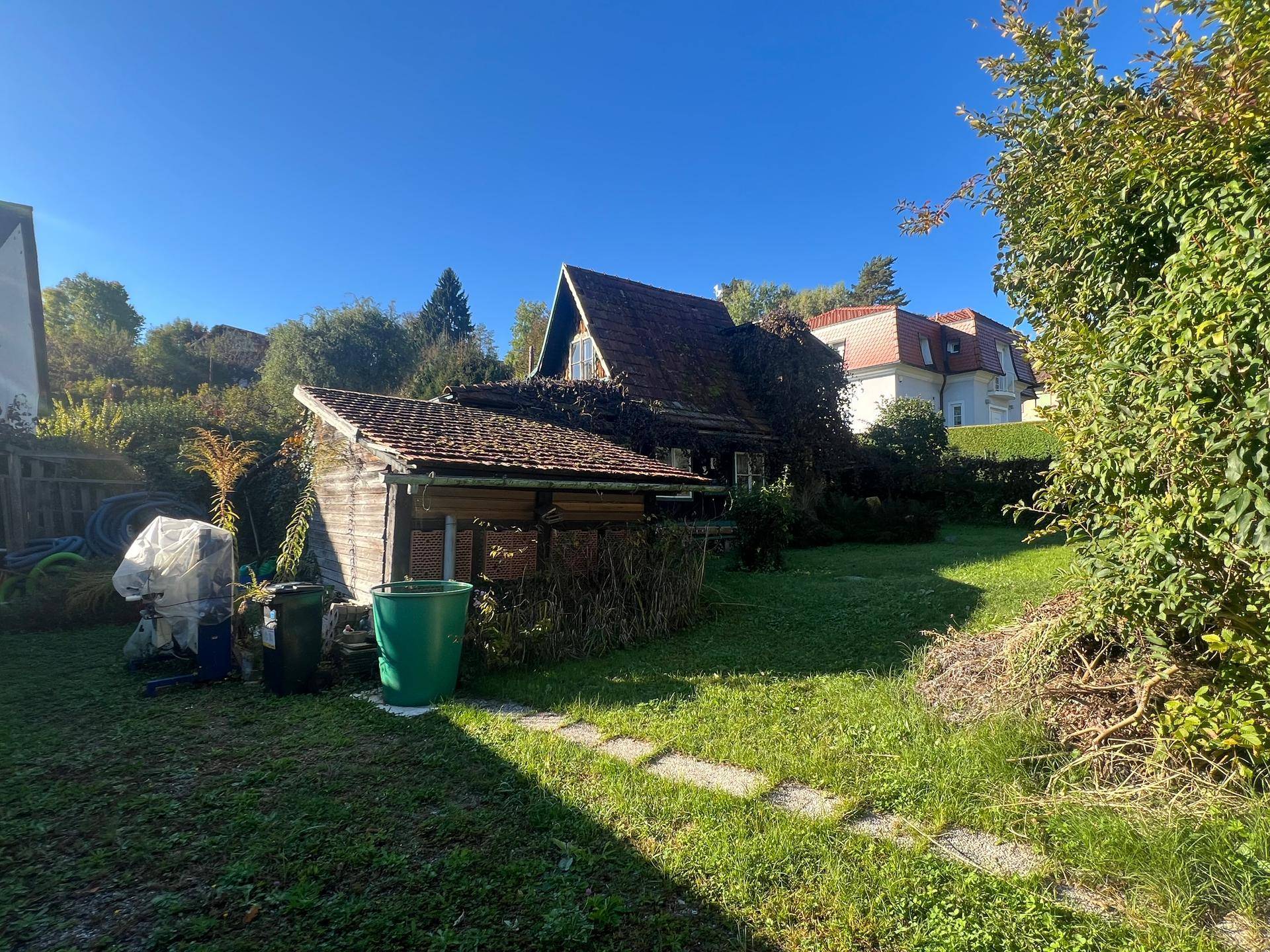 Rustikaler Holzschuppen mit Ziegeldach und Bewuchs, umgeben von einer grünen Rasenfläche und Bäumen.