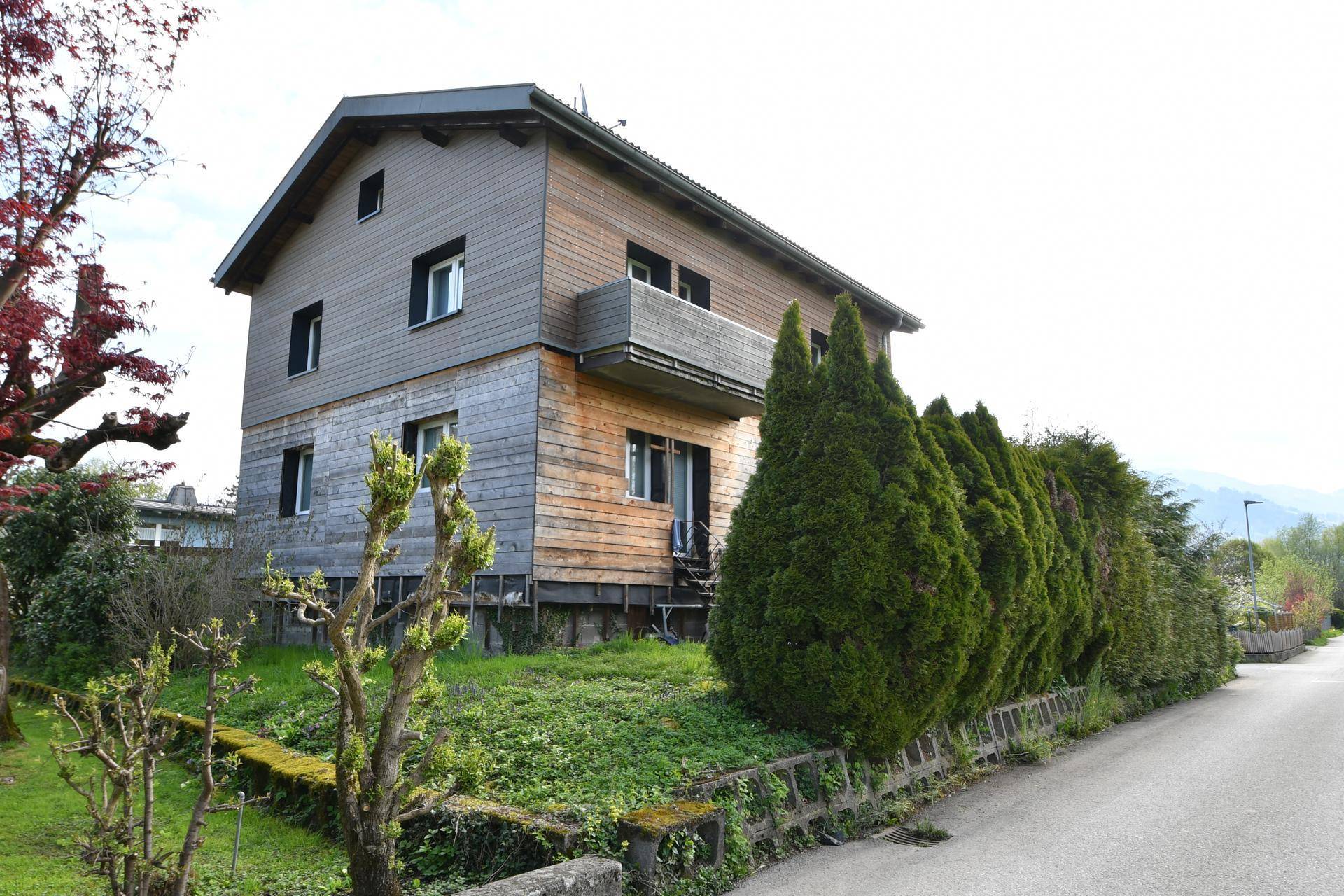 Wohnhaus mit Holzfassade und kleinem Balkon, umgeben von Hecken und Bäumen an einer Straße.