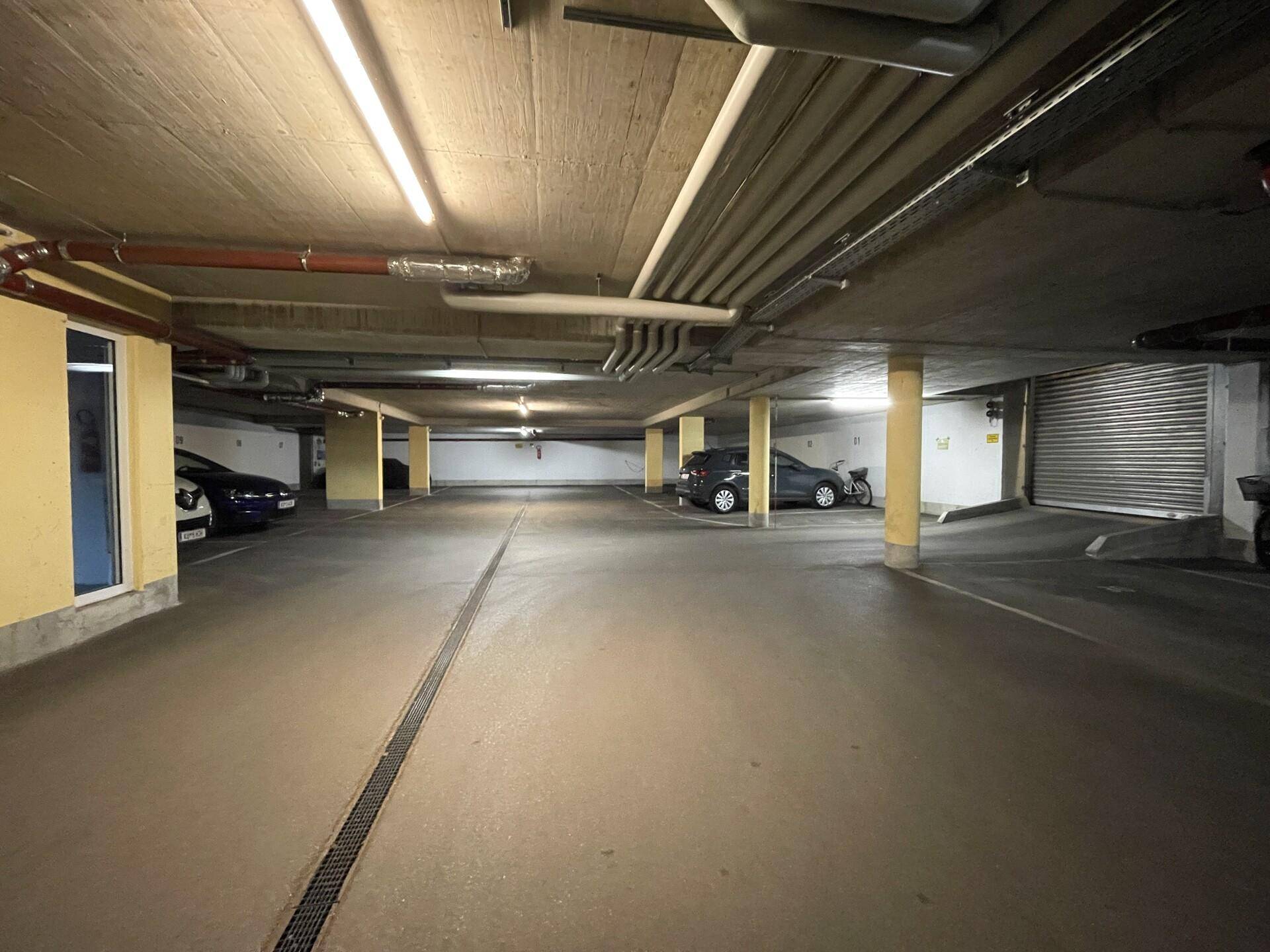 Geräumige Tiefgarage mit mehreren Stellplätzen, guter Beleuchtung und sichtbaren Rohren an der Decke.