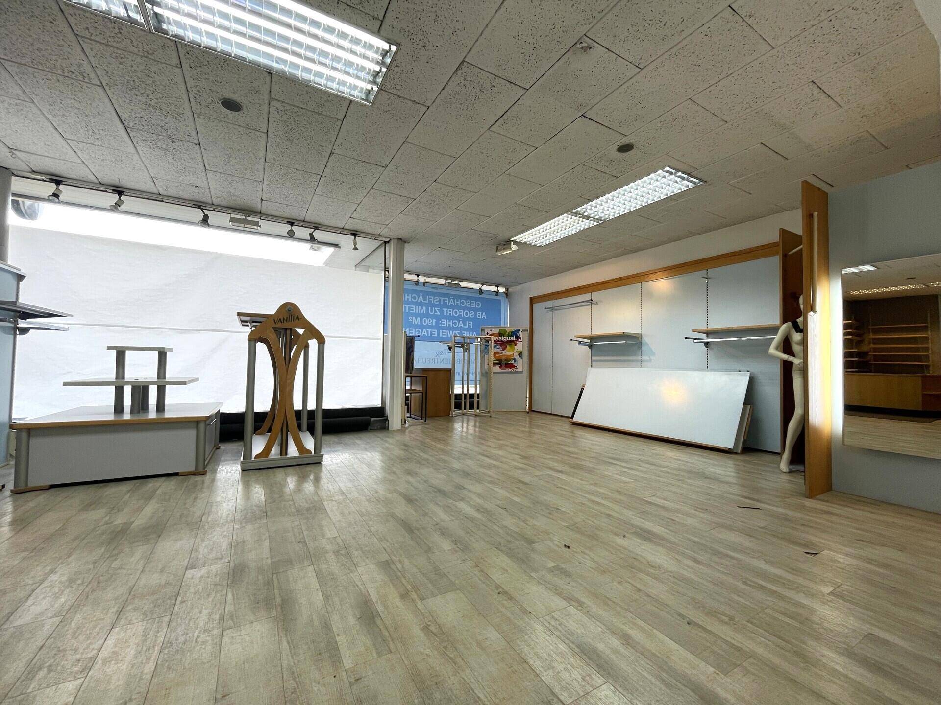 Innenansicht des Ladenlokals mit Schaufenstern, abgehängter Decke und einigen leeren Regalen und Schaufensterpuppen.