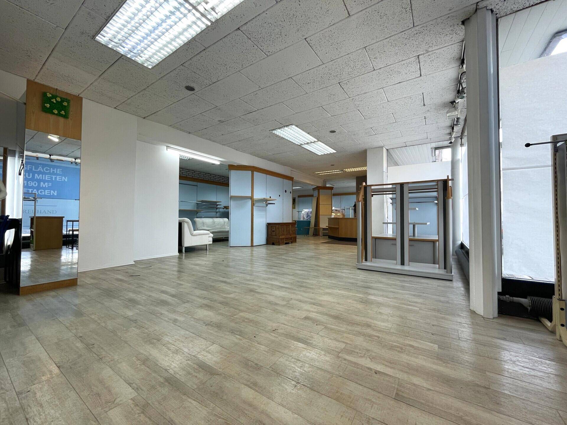 Geräumiger, leerstehender Ladenbereich mit hellem Holzboden, abgehängter Decke und großen Schaufenstern.