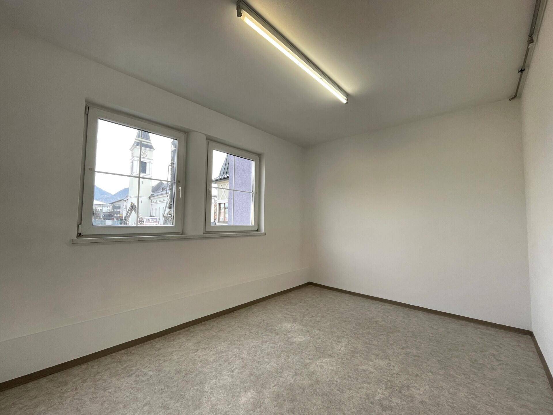 Heller Raum mit Linoleumboden, weißen Wänden und Fenstern, die einen Blick auf die Stadt und einen Kirchturm bieten.