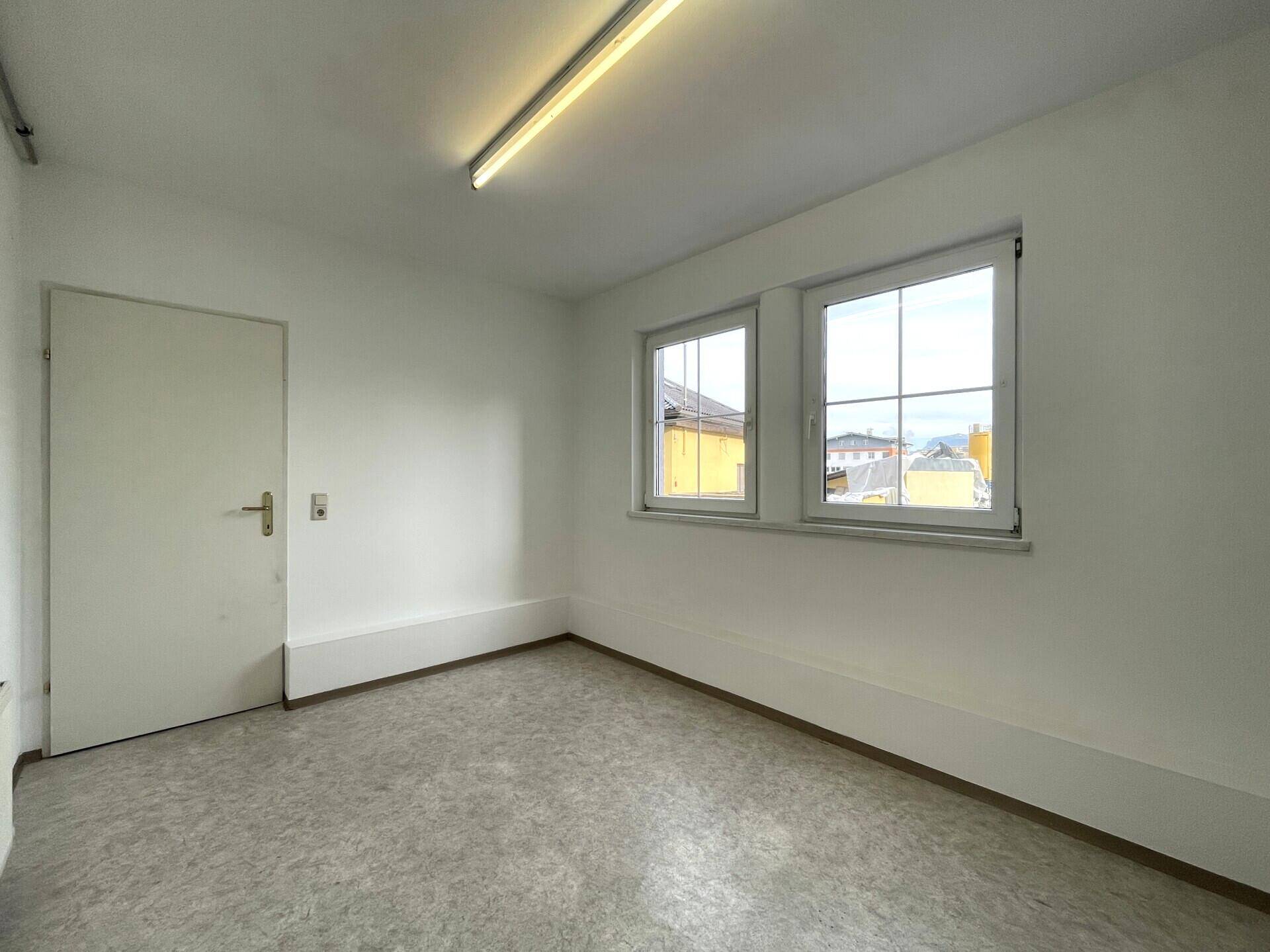 Kleiner, heller Raum mit Linoleumboden, weißen Wänden und zwei Fenstern mit Blick nach draußen.