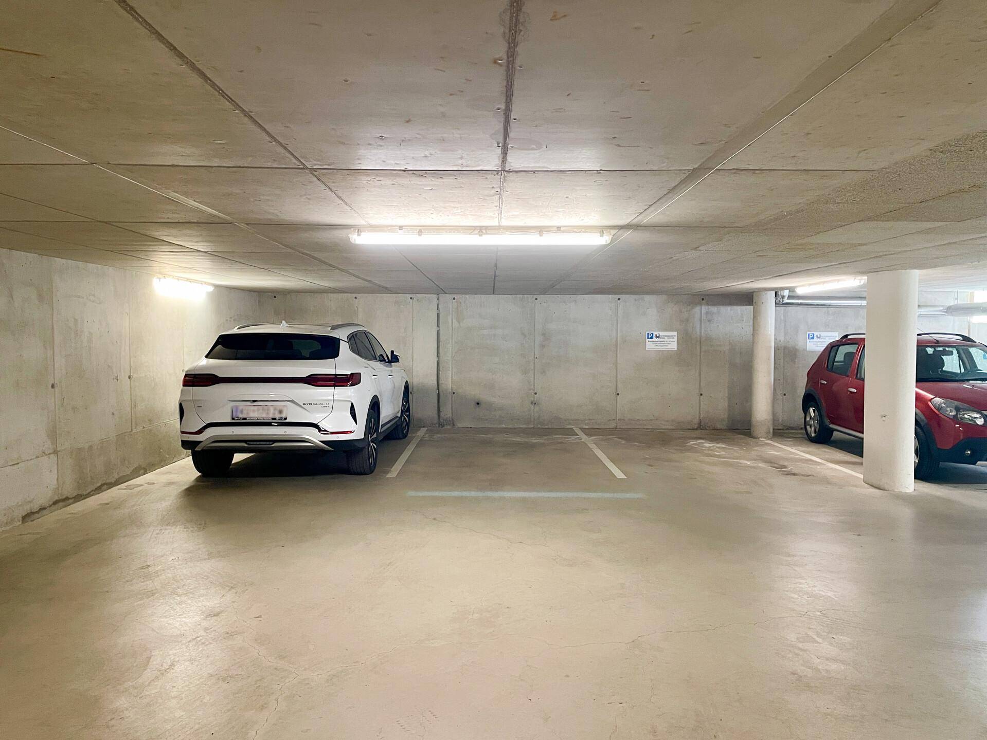 Helle Tiefgarage mit Betonwänden, Deckenbeleuchtung und markierten Parkplätzen.