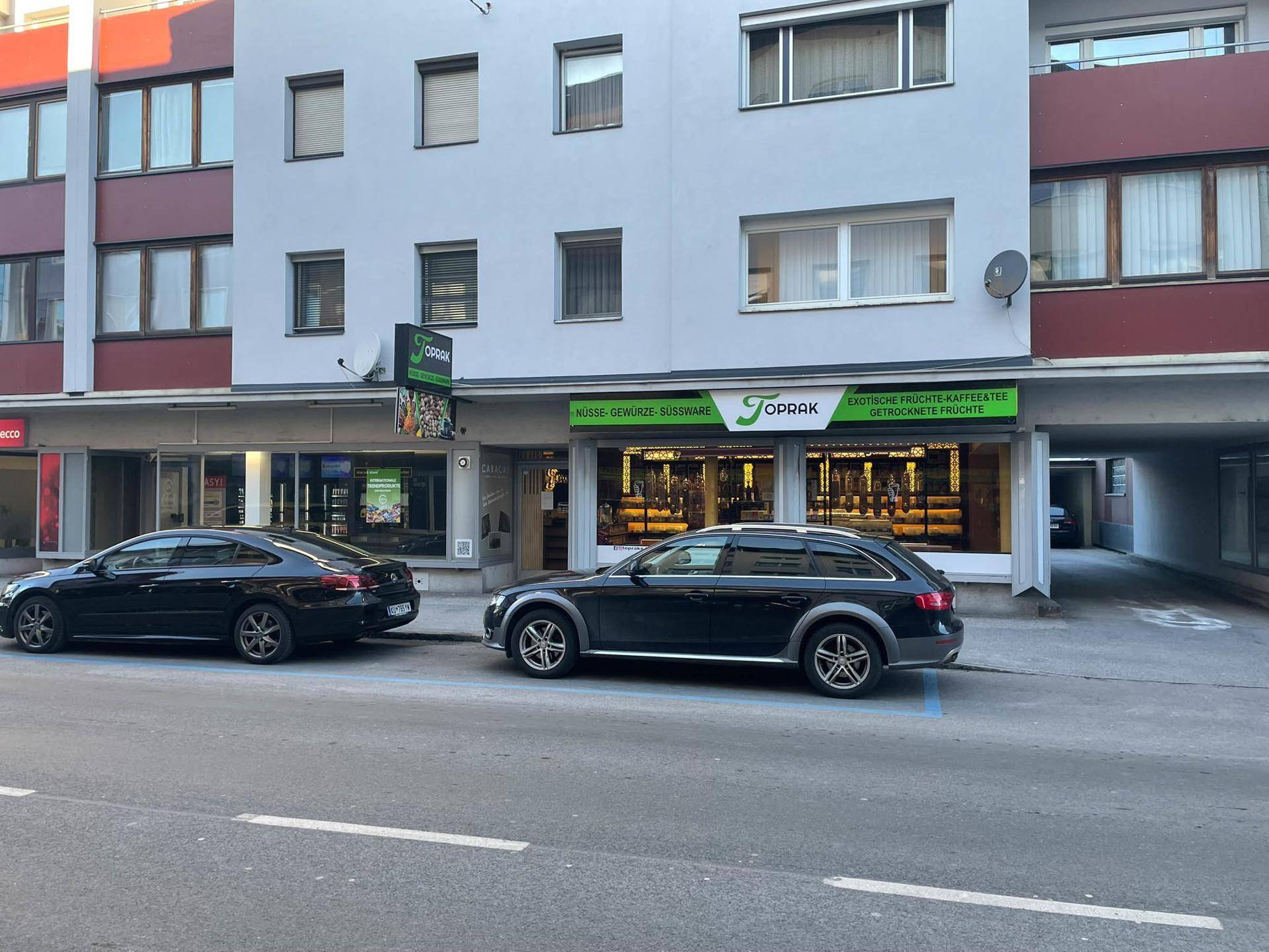 Gebäudefassade mit Erdgeschoss-Geschäften und darüber liegenden Wohnungen, Autos parken auf der Straße.