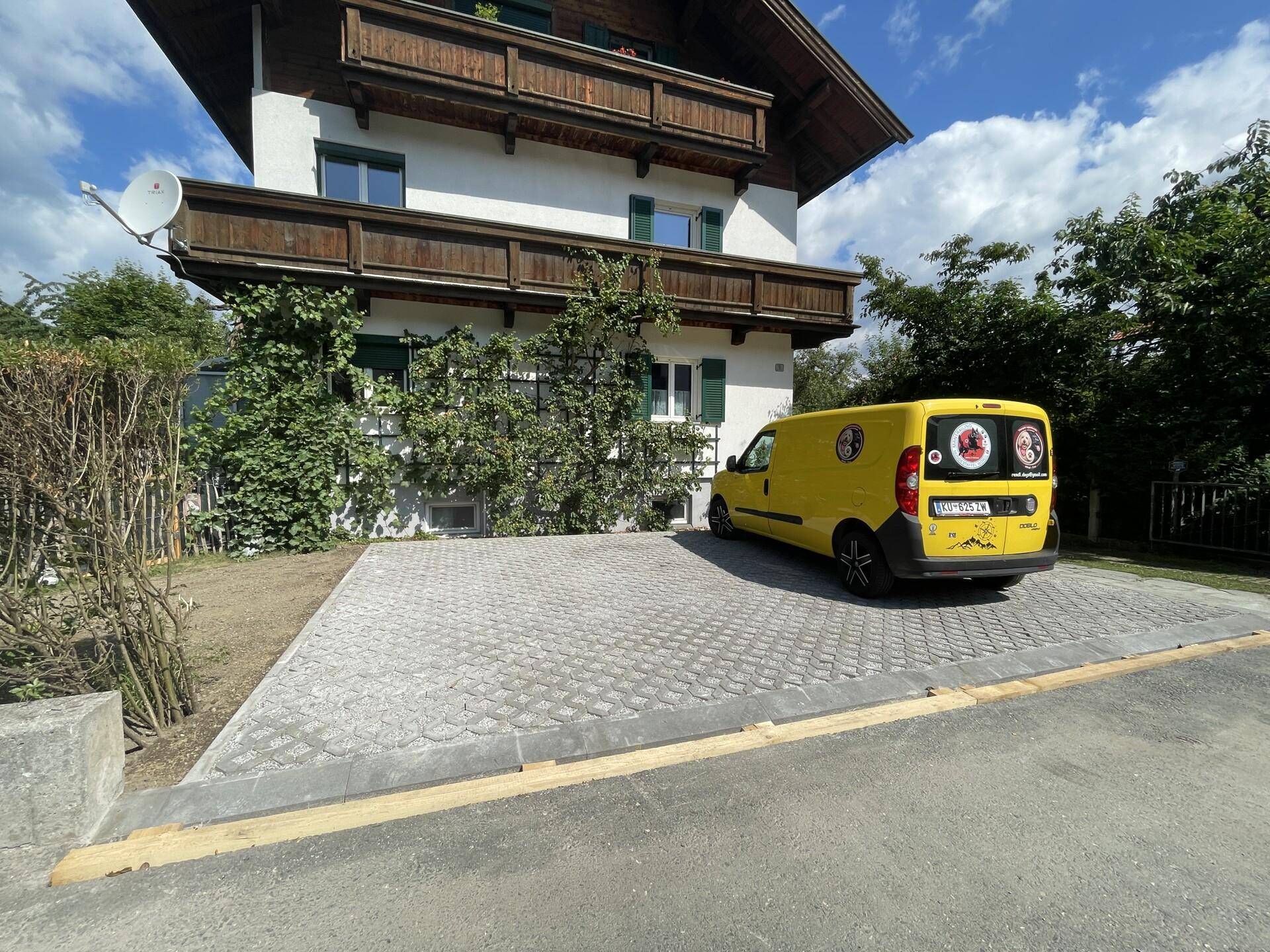 Traditionelles Wohnhaus mit Holzbalkonen und gepflastertem Parkplatz, auf dem ein gelber Lieferwagen steht.