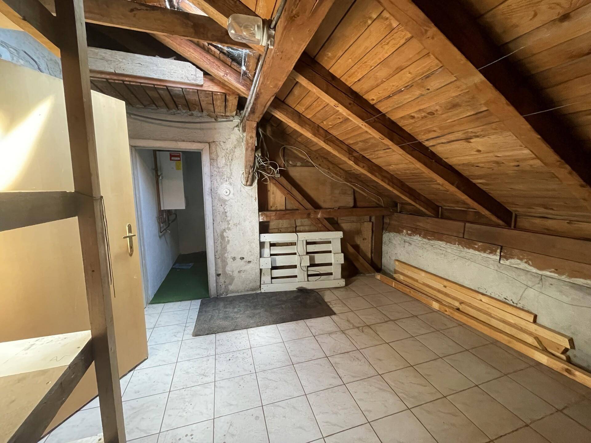Dachbodenraum mit Holzbalken, Fliesenboden und Zugang zu einem Nebenraum mit Warmwasserbereiter.