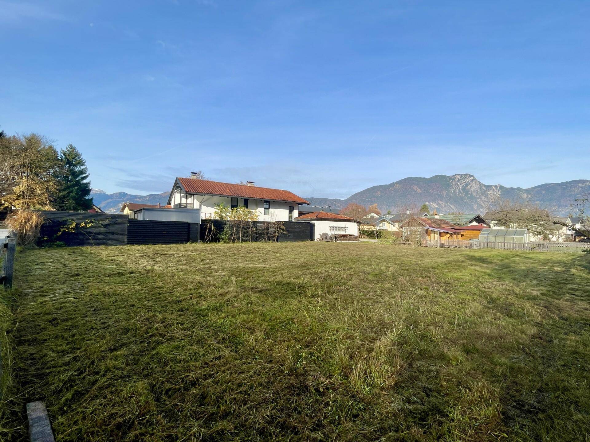 Weitläufige Grünfläche mit Wohnhäusern im Hintergrund und einer malerischen Bergkulisse.